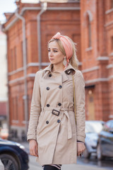 Stylish blonde girl in a beige raincoat in the city. Shoulder bag. The image of a modern woman in the city on the background of architecture