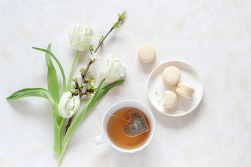Spring blossoms and a cup of tea with a heart shape tea bag and macaroons on a bright background, flat lay style with a copy space