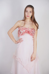 Fototapeta premium a charming young caucasian girl stands in a pink long prom dress with flower petals on her chest and poses on a white background in the Studio