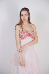 a charming young caucasian girl stands in a pink long prom dress with flower petals on her chest and poses on a white background in the Studio