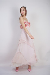 Fototapeta na wymiar a charming young caucasian girl stands in a pink long prom dress with flower petals on her chest and poses on a white background in the Studio