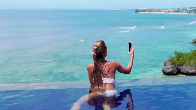 A young and beautiful girl in an open-air pool on a tropical island takes a smartphone camera landscape. Woman swimming in the pool overlooking the sea