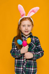 Obraz na płótnie Canvas Portrait of cute red-haired little child girl with Easter bunny ears holding colorful eggs on yellow background. Happy easter
