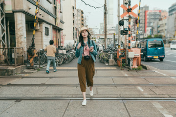 Fototapeta premium full length trendy young asian woman traveler walking outdoors holding mobile phone texting online message. beautiful female backpacker carrying professional camera while crossing railway osaka japan