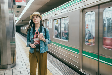 Naklejka premium Young woman typing on smart phone at tube station in osaka japan. girl travel backpacker looking up of road information indicator. beautiful japanese lady walking on platform and subway train leaving