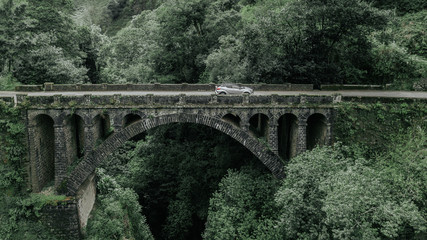 Fototapeta na wymiar Drone aerial view of a car crossing a stone bridge over a water stream in Cruzinhas, Faial, Madeira island, Portugal.
