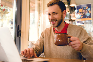 cheerful bearded blogger using laptop and holding cup of coffee in cafe