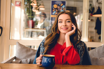 cheerful and attractive woman smiling while holding cup of coffee in cafe