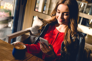 happy young woman holding cup of coffee and looking at smartphone in cafe