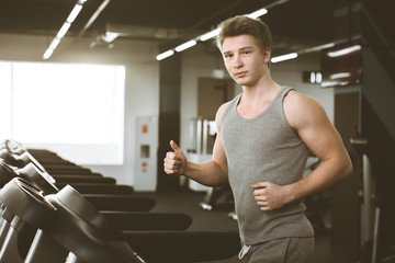 Young happy handsome man in grey sportswear in fitness club. Cardio workout, running on treadmill. Healthy lifestyle, guy and woman training in gym. Flare effect