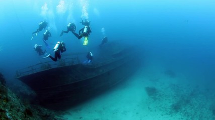 Group of divers swims near ship wreck underwater. Sunken ship covered in coral on Pacific Ocean seabed in Fiji.