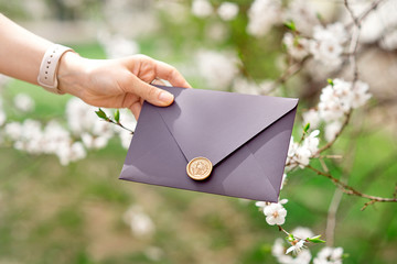 Close-up photo of female hands holding purple invitation envelope with wax seal, gift certificate, postcard, wedding invitation card on the background of blooming flowers