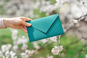 Close-up photo of female hands holding a green invitation envelope with a wax seal, a gift certificate, a postcard, a wedding invitation card on the background of blooming flowers