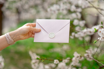 Close-up photo of female hands holding a pink invitation envelope with a wax seal, a gift certificate, a postcard, a wedding invitation card on the background of blooming flowers
