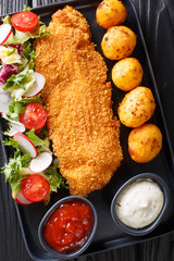 Portion of white fish fillet in breading with new potatoes and fresh vegetable salad close-up on the table. Vertical top view