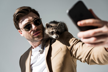 selective focus of handsome man with adorable raccoon on shoulder, taking selfie with smartphone on grey