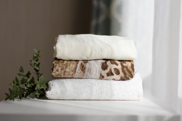 a stack of neatly folded clothes made of natural fabrics is decorated with a branch of a green plant on a white chest near the window