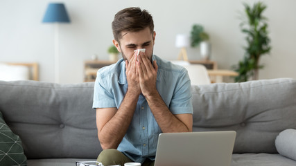 Sick man blowing nose, holding handkerchief, using laptop at home