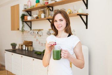 Smiling young woman drinking green smoothie juice in kitchen. Healthy Lifestyle.