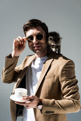 smiling man with furry raccoon on shoulder, holding coffee cup and looking at camera on grey