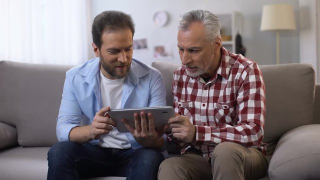 Retiree father and middle aged son scrolling tab, looking and smiling to camera
