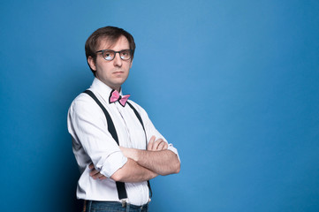 handsome serious man in shirt, suspender, pink bow tie and glasses standing and looking at camera with crossed arms on blue background with copy space