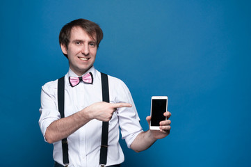 handsome man in shirt, suspender and pink bow tie standing and pointing with finger to smartphone with blank screen on blue background