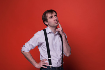 handsome dreamy man in shirt and black suspender standing with hand on hip, propping cheek with finger and looking away on orange background