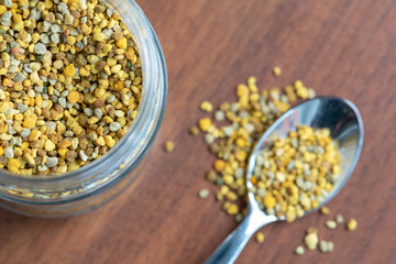 glass vase full of bee pollen granules with a teaspoon and pollen scattered on the table, seen from above