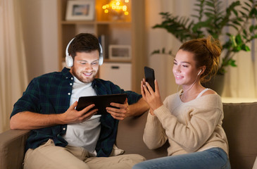 leisure, technology and people concept - happy couple with tablet computer, headphones and smartphone listening to music at home in evening