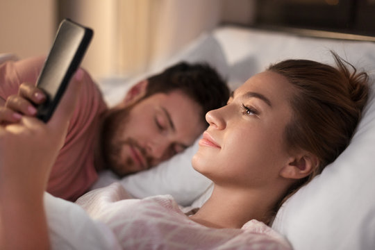 technology, internet addiction and cheat concept - woman using smartphone at night while her boyfriend is sleeping