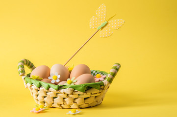 Close up. Easter still life. Rustic eggs in a wicker basket. The decor of the felt flowers and butterfly of pastel colors. Yellow background. Copy space.