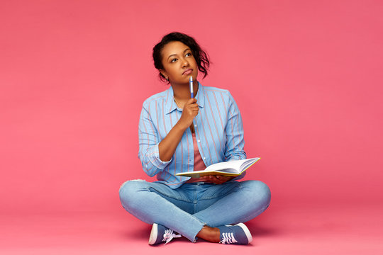 education, high school and inspiration concept - african american young student woman with pen and diary or notebook sitting on floor and thinking over pink background