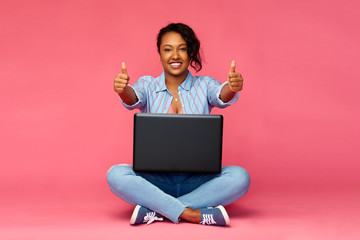 people and technology concept - happy african american young woman with laptop computer sitting on floor and showing thumbs up over pink background