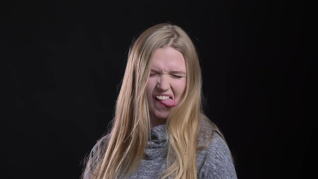 Portrait of young blonde straight-haired model showing funny emotions into camera on black background.