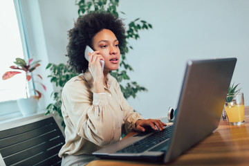 Smiling young African female entrepreneur sitting at a desk in her home office working online with...