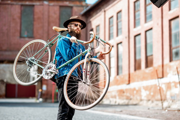 Lifestyle portrait of a bearded hipster dressed stylishly with hat and jacket carrying his retro bicycle on the urban background