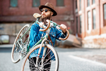 Lifestyle portrait of a bearded hipster dressed stylishly with hat and jacket carrying his retro bicycle on the urban background