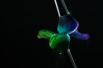 Cosmetic shades of different colors, blue and green, fly away from two make-up brushes creating a fancy pattern on a black background.