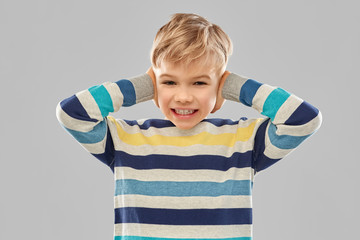 noise, stress and people concept - stressed little boy in striped pullover closing ears by hands over grey background