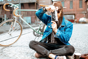 Bearded hipster dressed stylishly with hat and jacket sitting with photo camera and retro bicycle on the street outdoors