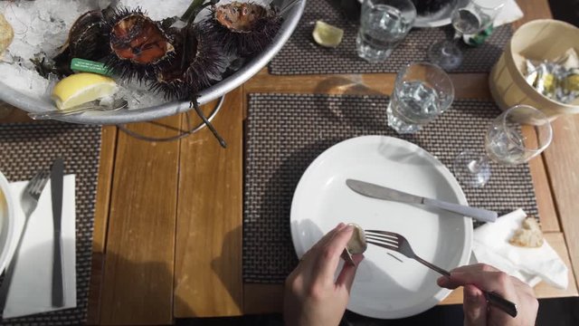 Man grabs a mussel with his fingers from an ice bowl and tries to remove it from its shell with a fork at an outdoor seafood restaurant by a sunny day