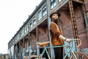 Fototapeta na wymiar Lifestyle portrait of a bearded hipster dressed stylishly walking with retro bicycle on the industrial urban background