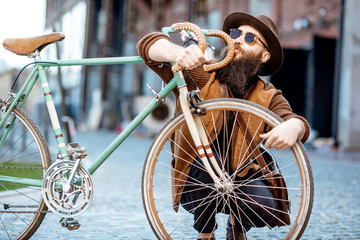 Portrait of a bearded hipster dressed stylishly with hat hugging his retro bicycle outdoors on the urban background