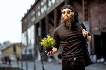 Portrait of a serious bearded man as a killer dressed in black tight clothes holding flower pot and...