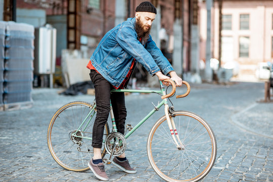 Lifestyle portrait of a bearded hipster dressed stylishly with hat and jacket standing with retro bicycle on the industrial urban background