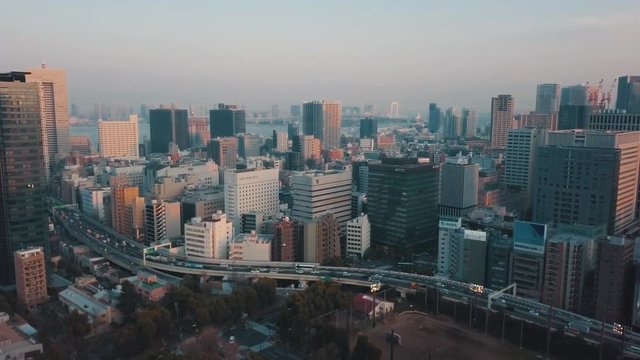 Drone panning up above the Tokyo city skyline at sunset over a bustling highway bridge, tall skyscrapers and orange light reflections with hazy skies