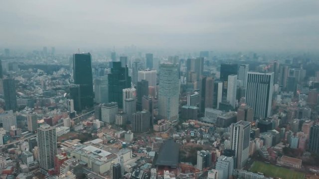 Aerial drone panning above the Tokyo city skyline in Japan on an overcast day with hazy smog filled skies and grey storm clouds above roads, skyscrapers and a sports field