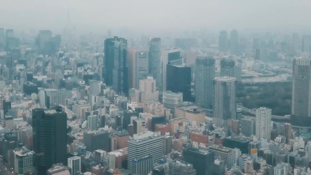 Aerial drone panning up above the Tokyo city skyline in Japan on an overcast day with hazy smog filled skies and grey storm clouds above busy roads and skyscrapers