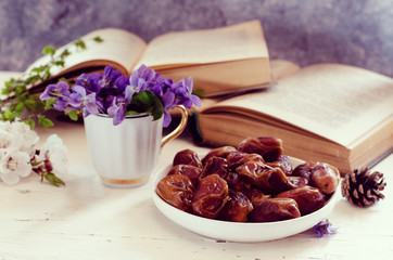Still life with violet in white cup, old books and date fruits in a plate. Romantic floral background.